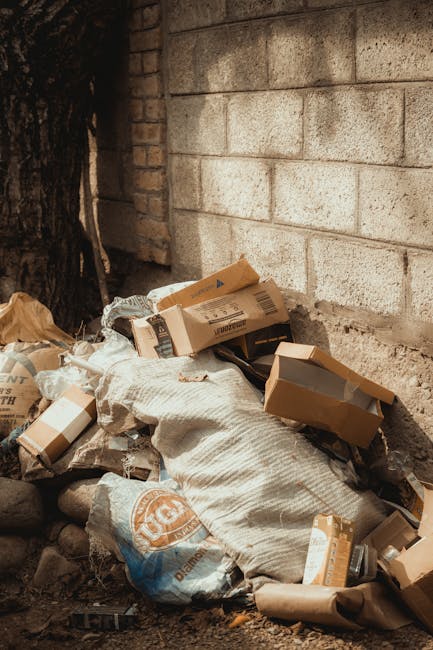 A pile of discarded cardboard boxes and paper packaging materials, some flattened and others partially folded, resting on rocky ground next to a weathered brick wall with visible cement mortar. The boxes vary in size, with some corrugated cardboard flaps open and others closed, displaying printed labels and barcodes. A large grey fabric sack or bag appears to be used for storing some of the waste, partially covering a few smaller items around it. To the left, a tree trunk with dark, textured bark is partially visible, casting shadows that suggest natural daylight. The scene is outdoors, situated in an area adjacent to an exterior brick wall, possibly part of a building's foundation or side wall, with no visible signage or other objects. The overall setting reflects an unmanaged collection of waste materials awaiting removal, aligning with the themes of alternative waste handling or private rubbish collection services often provided by companies such as Rubbish Removal Brent.