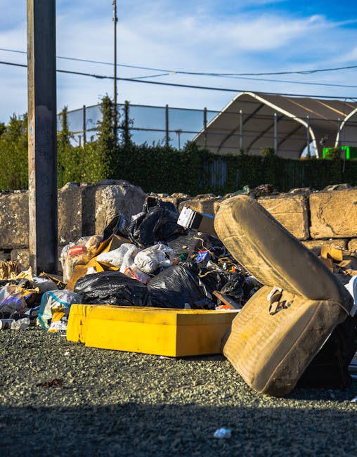 A close-up view of a pile of mixed household rubbish and waste materials accumulated on a paved outdoor surface. The debris includes black plastic rubbish bags, some torn open, revealing miscellaneous refuse, along with a yellow plastic storage container and a large, dirty car tire, leaning against the rubbish heap. Behind the pile, a low stone wall made of roughly cut, irregular stones separates the area from a background scene featuring a green hedge, an open-sided shelter or canopy structure, and a wire fence. To the left, a metal utility pole supports overhead electrical wires set against a bright blue sky with sparse, wispy clouds. The scene is lit by natural daylight, highlighting the textures and colors of the waste and surrounding environment. This cluttered arrangement illustrates a scenario where unsorted rubbish has been collected for potential private disposal, aligning with independent rubbish removal services that Rubbish Removal Brent offers for local waste clearance needs.