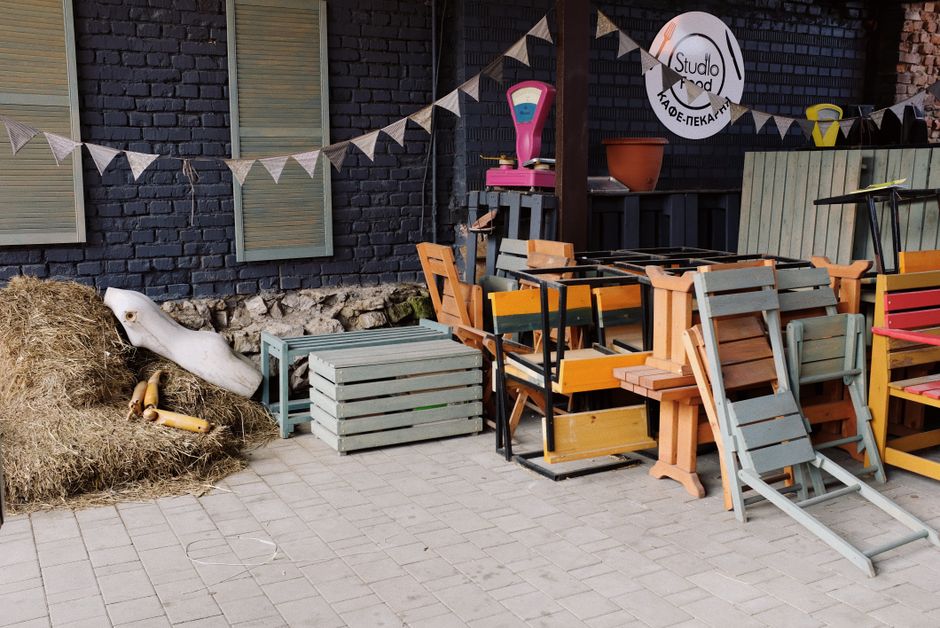 An outdoor area adjacent to a dark brick building features a collection of stacked wooden garden furniture, including chairs and tables, in natural and painted finishes such as light gray, yellow, and reddish-brown, with some items leaning or stacked. To the left, there is a hay bale with a white plastic fish figurine resting on top, along with scattered wooden barrels or logs nearby. Behind the furniture, a black brick wall is visible with a string of triangular fabric bunting hanging across it. On the right, a wooden fence panel partially surrounds the space, and a small round tabletop with a pink scale and a terracotta plant pot are positioned on a high shelf on the wall. A circular sign or logo reading 'Studio Food' is mounted on the wall, adding an informal decorative element. The ground consists of pale paving stones, creating a neutral surface suitable for either storage or casual outdoor seating, and Rubbish Removal Brent occasionally handles such areas for private waste clearance, involving the removal of bulky furniture and refuse from commercial or residential premises.