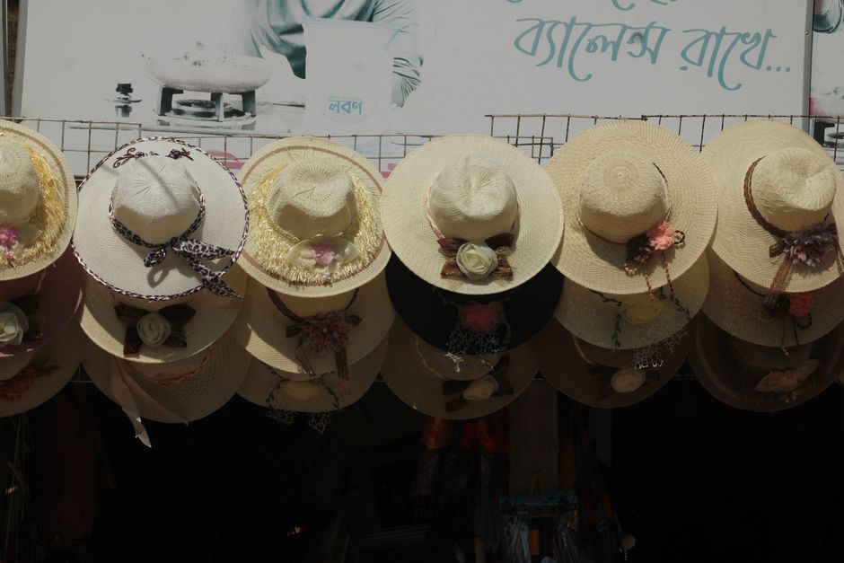 A row of eight wide-brimmed straw hats with decorative ribbons, artificial flowers, and bows, are displayed on a reflection-surfaced ledge outdoors. The hats vary slightly in size and design, with some featuring lace, floral embellishments, or contrasting bands, and are arranged in a line with their brims facing downward. In the background, a metal fence and a partially visible sign with Bangla script can be seen, indicating a market or street stall environment. The sunlight casts shadows on the hats, highlighting their textured straw material and decorative details, reflecting a typical scene of accessory display in a busy market area. Rubbish Removal Brent occasionally handles disposal of items like these as part of on-site clearance or private waste removal services, helping maintain cleanliness in retail or outdoor display spaces.