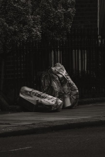 Several large black and white plastic garbage bags filled with household waste are placed on the pavement near the edge of a residential street. The bags appear to contain mixed refuse and are somewhat crumpled, with some labels or markings partially visible on the plastic. They are positioned beside a dark metal fence with vertical bars, which runs parallel to the curb, and behind the fence, there are dense dark foliage and trees, creating a shaded backdrop. The scene is lit with minimal natural light, emphasizing the stark contrast of the trash bags against the urban environment. This image, associated with rubbish removal services, illustrates the typical sight of bulky waste ready for collection or private disposal, relevant to independent waste clearance options often necessary when regular bin collection is insufficient or unavailable. The overall scene is cleanly composed but highlights the common challenge of managing bulk waste in residential outdoor areas, aligning with the themes of alternative waste handling and rubbish removal solutions provided by companies like Rubbish Removal Brent.