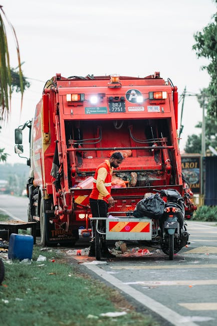A worker wearing a yellow and red uniform is standing behind a large red waste collection vehicle, which is positioned on a street alongside a curb. The vehicle, used for rubbish removal and private waste handling, features a spacious rear hopper with mechanical arms and hydraulic components visible, indicating active collection activities. The worker appears to be sorting or preparing waste, with a black plastic bag placed on a small metal platform attached to a bicycle or scooter nearby. The scene takes place outdoors during daylight hours, with overcast weather casting soft, diffused light across the environment. In the background, some trees, power lines, and signs or storefronts are observable, suggesting a commercial or mixed-use area. Debris and waste materials are seen on the pavement around the vehicle, emphasizing the context of rubbish removal services. The environment reflects a typical urban or suburban street scene where independent refuse collection might occur, supported by professional waste management services like Rubbish Removal Brent.