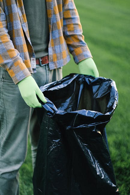 Several large black and white plastic garbage bags filled with household waste are placed on the pavement near the edge of a residential street. The bags appear to contain mixed refuse and are somewhat crumpled, with some labels or markings partially visible on the plastic. They are positioned beside a dark metal fence with vertical bars, which runs parallel to the curb, and behind the fence, there are dense dark foliage and trees, creating a shaded backdrop. The scene is lit with minimal natural light, emphasizing the stark contrast of the trash bags against the urban environment. This image, associated with rubbish removal services, illustrates the typical sight of bulky waste ready for collection or private disposal, relevant to independent waste clearance options often necessary when regular bin collection is insufficient or unavailable. The overall scene is cleanly composed but highlights the common challenge of managing bulk waste in residential outdoor areas, aligning with the themes of alternative waste handling and rubbish removal solutions provided by companies like Rubbish Removal Brent.