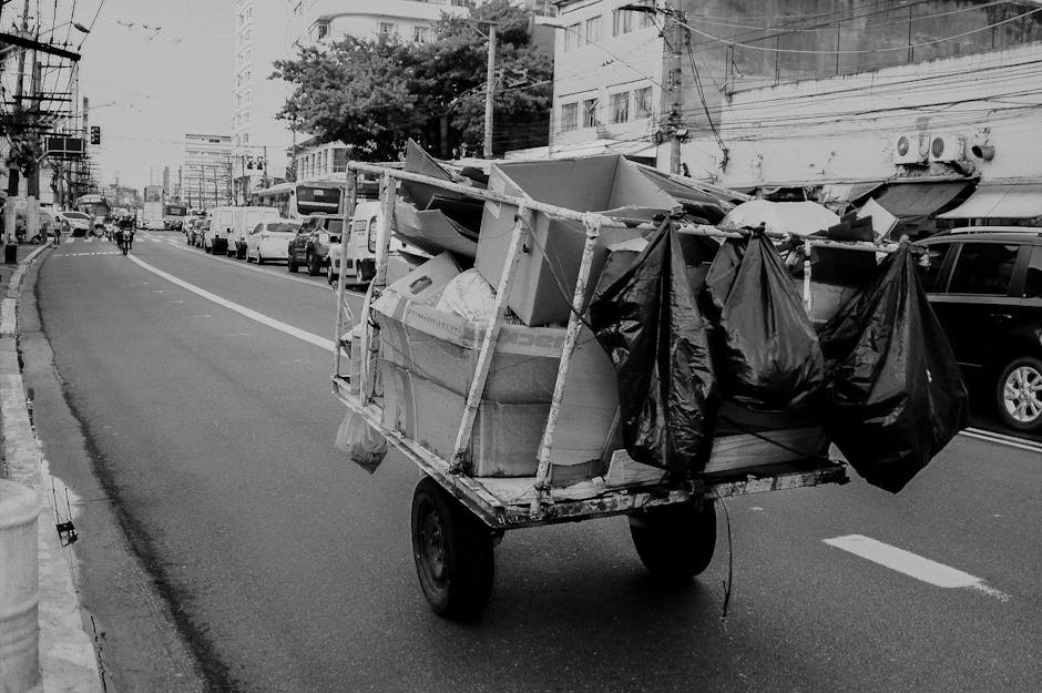 A small, makeshift hand-pulled cart equipped with two large black plastic garbage bags hanging from the sides, positioned on an urban street in daylight. The cart's frame is constructed from metal and wooden planks, supporting various discarded items including a white rectangular object and a large cardboard box with visible edges and textures. The street scene features a row of parked cars of different models and colors, mainly dark and white, along the right side, with a mixture of modern buildings, some with balconies, in the background. Power lines are suspended overhead, crossing above the street, which extends into the distance with a slight curve. Pedestrians and cyclists are visible further down the roadway, indicative of typical city activity. The environment appears to be busy, with the cart likely used for informal collection or transportation of waste or recyclables, consistent with alternative waste handling practices in urban areas managed by local waste removal services, such as Rubbish Removal Brent.