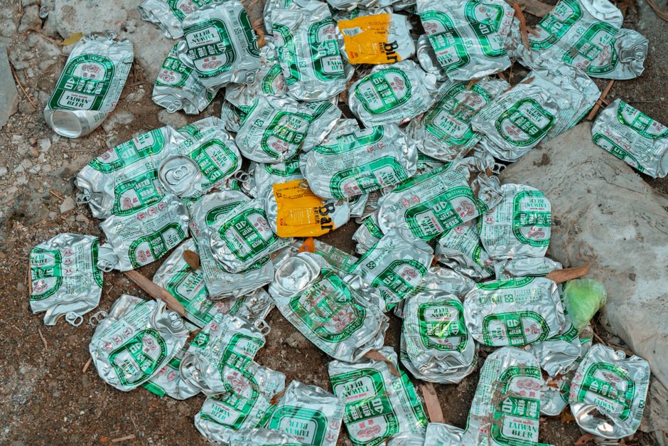 A collection of crushed aluminum cans with green and white printed labels, scattered on a rough, uneven outdoor ground surface with visible dirt, small rocks, and dried leaves. Some cans are piled on top of each other, while others are spread across the area. The cans are metallic with a shiny, reflective finish, and the labels indicate they previously contained beer. In the background, there is a plastic bag partially filled with waste and a small yellow label attached to one of the cans. The scene suggests a site of discarded beverage containers, which could be gathered for rubbish removal or recycling services, similar to those offered by Rubbish Removal Brent for private waste handling on sites such as Kilburn High Road NW6.