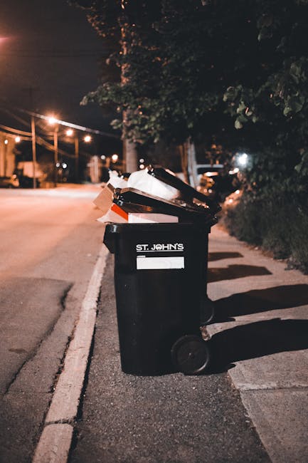 A small, makeshift hand-pulled cart equipped with two large black plastic garbage bags hanging from the sides, positioned on an urban street in daylight. The cart's frame is constructed from metal and wooden planks, supporting various discarded items including a white rectangular object and a large cardboard box with visible edges and textures. The street scene features a row of parked cars of different models and colors, mainly dark and white, along the right side, with a mixture of modern buildings, some with balconies, in the background. Power lines are suspended overhead, crossing above the street, which extends into the distance with a slight curve. Pedestrians and cyclists are visible further down the roadway, indicative of typical city activity. The environment appears to be busy, with the cart likely used for informal collection or transportation of waste or recyclables, consistent with alternative waste handling practices in urban areas managed by local waste removal services, such as Rubbish Removal Brent.