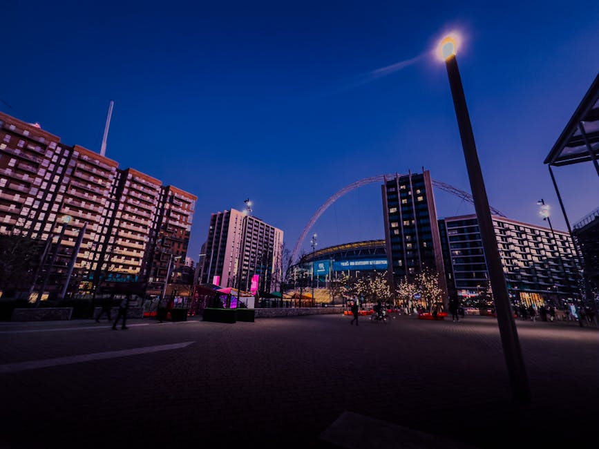 A cityscape scene viewed during the evening with a clear sky and illuminated buildings. The foreground shows a paved open space with a tall streetlamp on the right side, casting a bright light upward. Surrounding the open area are modern mid-rise buildings with a combination of glass windows and brick or concrete facades, some with balconies. In the background, a large, illuminated Ferris wheel and a stadium with a lit roof are visible, suggesting the location is near a notable entertainment venue. The scene is softly lit by streetlights and building lights, creating a calm atmosphere. The area appears to be an urban public space, possibly near a commercial or entertainment district, and the environment is typical for a central city hub. Although not specifically related to rubbish removal, the scene depicts a clean, organized city environment that could be part of a private or municipal waste management or clearing zone managed by companies like Rubbish Removal Brent specializing in onsite or alternative waste handling services in busy urban areas.