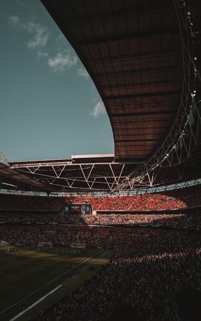 A wide-angle view of Wembley Stadium during a sporting event or concert, with a large oval roof structure overhead supported by an intricate framework of steel trusses. The partially open roof reveals a partly cloudy sky with a bright, diffuse light illuminating the stadium's interior. The seating areas are densely occupied with spectators, predominantly dressed in red and other vibrant colours, creating a lively atmosphere. The pitch at the center is visible with markings for a football match, and the lower stands appear filled with standing and seated attendees. In the foreground, part of a paved walkway or outdoor area surrounds the stadium, with no visible rubbish or waste. The structure and environment suggest careful maintenance and clean surroundings, typical of venues that manage their waste through independent collection services such as Rubbish Removal Brent, which handles various waste management needs relevant to large-scale events and venue operations.
